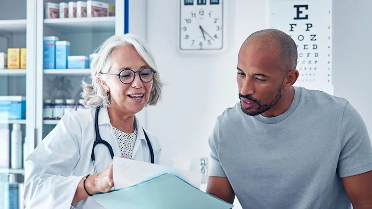 A doctor and a patient sitting together in a medical exam room, reviewing documents and discussing information in a folder. The setting includes medical cabinets, charts, and equipment, creating a calm, professional healthcare environment.