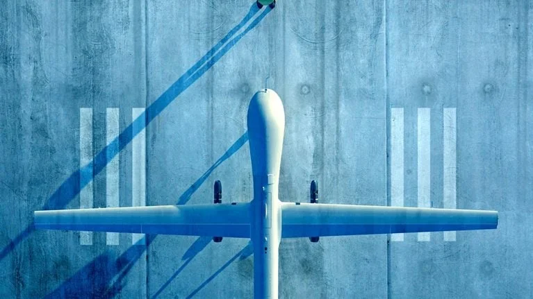 An overhead view shows a large unmanned aircraft parked on a concrete runway, its long wings stretching symmetrically across the frame. A small person nearby casts a long shadow, emphasizing the scale of the scene.