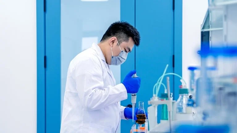 A lab researcher wearing a face mask, gloves, and a white coat is using a pipette to transfer liquid into a flask on a laboratory bench. The scene shows a clean, modern lab environment with scientific equipment and containers arranged for experimental work.